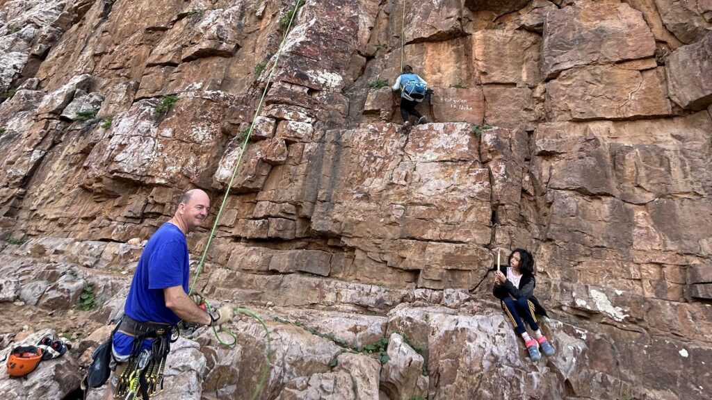 Parent belaying a climber on a tall rock wall while a child sits at the base of the crag in Morocco